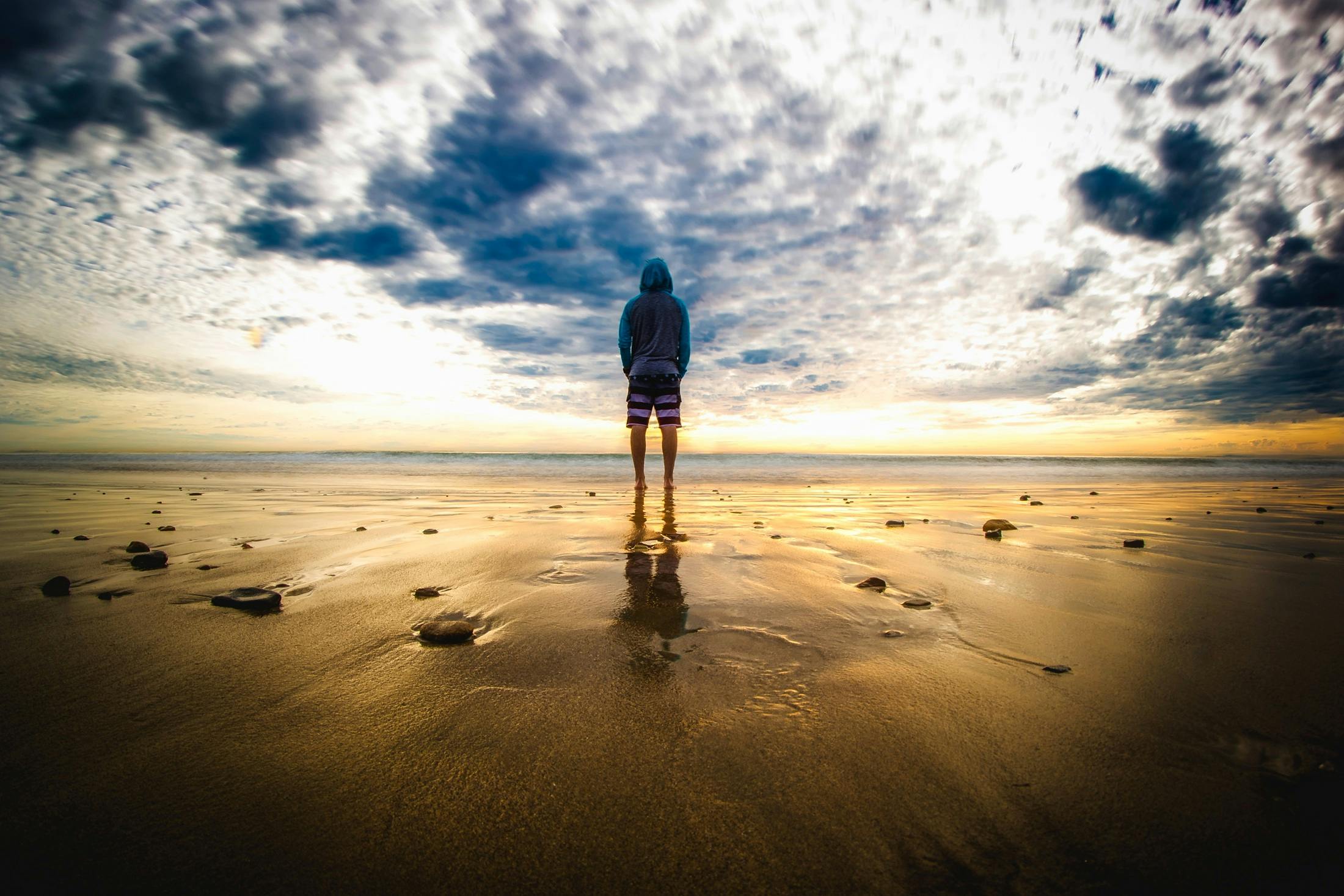 Prestations A lone figure stands on a tranquil beach during a vivid sunset, reflecting on the wet sand.